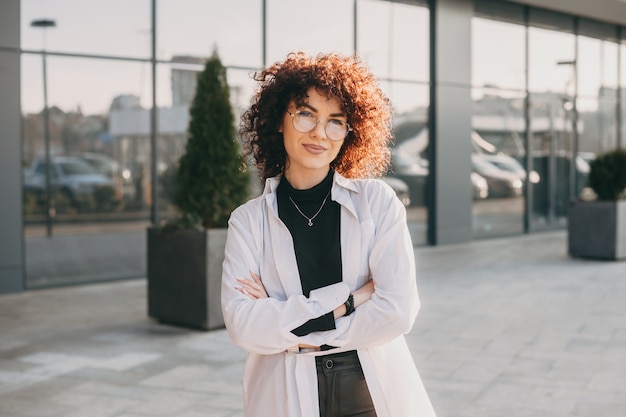 Charmante blanke vrouw met krullend haar kijkt door een bril terwijl ze buiten poseren met gekruiste handen