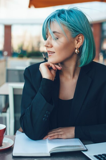 Charmante blauwharige vrouw poseren in een coffeeshop tijdens het schrijven van enkele ideeën op een notitieblok