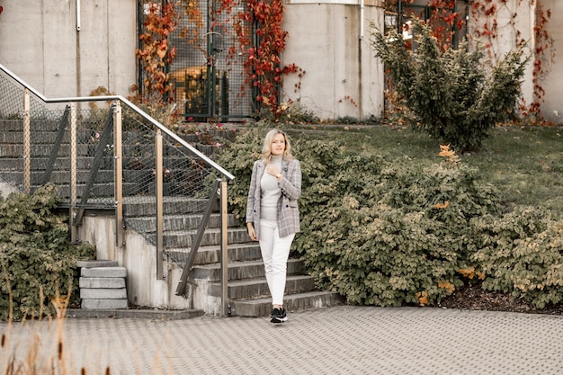 Charmante blonde met lang haar in jas en broek staat in de buurt van trappen op straatmode in de stad