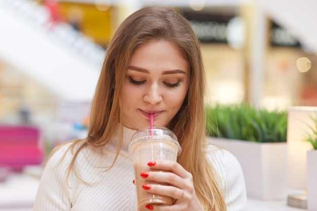 Charmante blonde vrouw drinkt cocktail, poseert in café, draagt witte casual trui, heeft rode manicure, kijkt neer op haar drankje, houdt van smaak, geniet van uitgaan