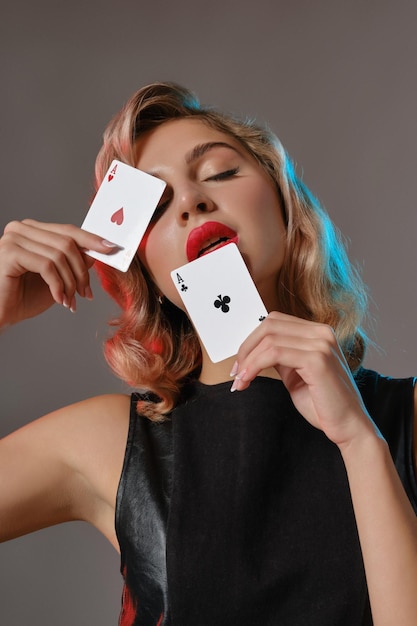 Charmante blonde vrouw met lichte make-up in zwarte stijlvolle jurk toont twee speelkaarten en poseren tegen grijze studio achtergrond Concept van gokken entertainment poker casino close-up