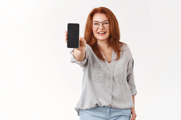 Charmante elegante roodharige zakenvrouw van middelbare leeftijd, moeder draagt een bril, strek de arm uit, houd de smartphone vast, toon telefoonschermweergave, promoot de toepassing witte muur