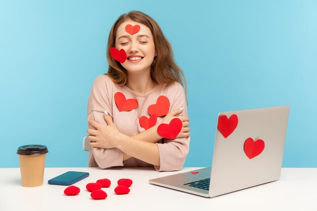 Charmante gelukkige jonge vrouw met vriendelijke glimlach zittend op het kantoor van de werkplek, allemaal bedekt met sticker liefdesharten, zichzelf omhelzend, genietend van Valentijnsdaggroeten. indoor studio-opname, geïsoleerd