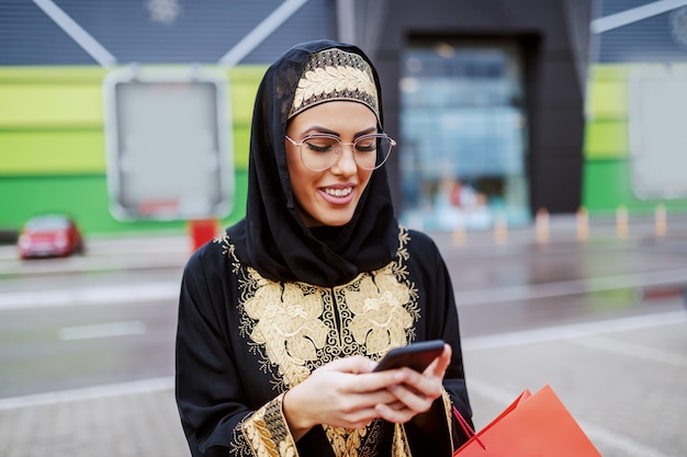 Charmante glimlachende Moslimvrouw in traditionele slijtage staande voor winkelcentrum met winkelen slecht in handen en nieuwe slimme telefoon uitproberen. Duizendjarige generatie. Diversiteit concept.