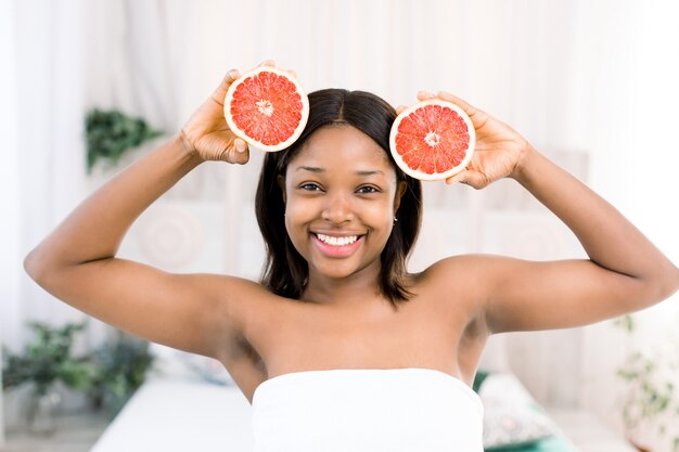 Charmante jonge afrikaanse meisje met plakjes grapefruit voor haar gezicht. foto van glimlachende afrikaanse amerikaanse vrouw die op lichte achtergrond wordt geïsoleerd. schoonheid huidverzorging concept