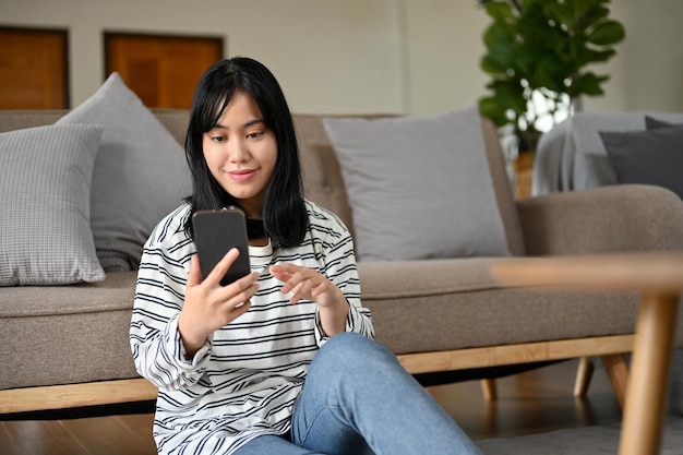 Charmante jonge aziatische vrouw die haar smartphone gebruikt terwijl ze ontspant in haar woonkamer