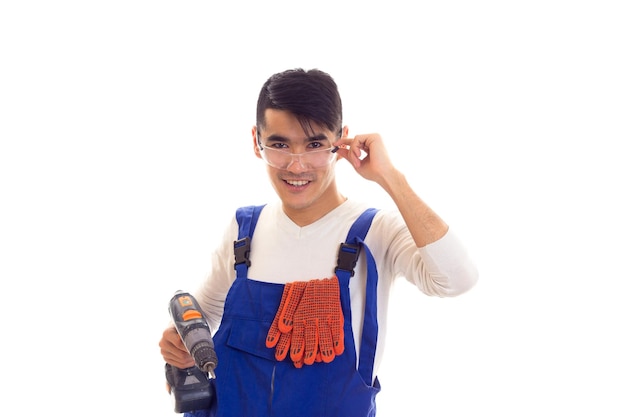 Charmante jonge man met donker haar in wit overhemd en blauwe overall met oranje handschoenen en beschermende bril met elektrische schroevendraaier op witte achtergrond in studio.