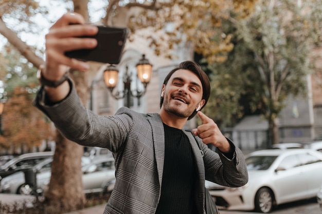 Charmante jonge man met donkerbruin haar en borstelharen, donkere trendy outfit dragend, selfie makend terwijl hij poseert op herfststraat bij daglicht