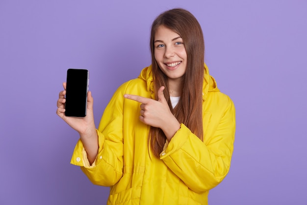 Charmante jonge mooie vrouw poseren geïsoleerd over lila achtergrond, met slimme telefoon, met lege display, wijzend op apparaat met haar vingers, glimlachend, kopieer ruimte voor reclame.