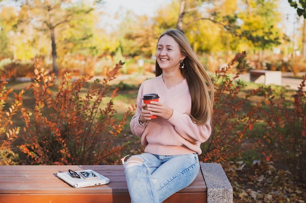 Charmante jonge vrouw drinkt koffie zittend op een bankje in een prachtig herfstpark