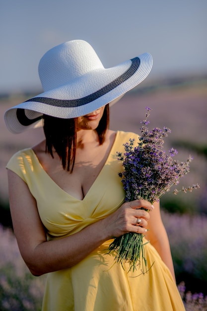 Charmante jonge vrouw in bloeiende lavendelvelden Achteraanzicht van mooie dame met een wuivende gele jurkhoed Lavendel etherische olie met verse lavendelbloemen aromatherapie spa massageconcept