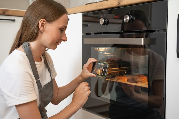 Charmante jonge vrouw in een grijs koksschort zet koekjes in de oven en maakt foto's op haar telefoon