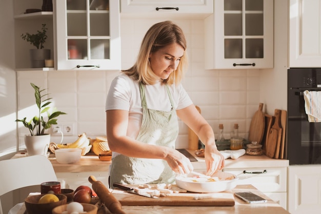 Charmante jonge vrouw in koksschort bereidt taart thuis in de keuken Gezellig interieur