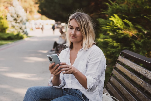 Charmante jonge vrouw in vrijetijdskleding zit op een bankje in het park en gebruikt een mobiele telefoon om te communiceren