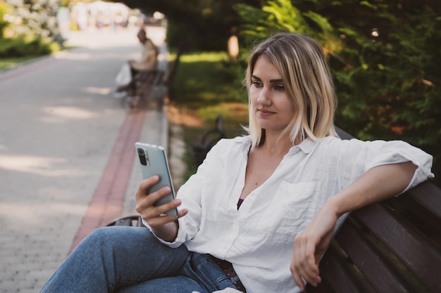 Charmante jonge vrouw in vrijetijdskleding zit op een bankje in het park en gebruikt een mobiele telefoon om te communiceren