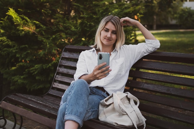 Charmante jonge vrouw in vrijetijdskleding zit op een bankje in het park en gebruikt een mobiele telefoon om te communiceren