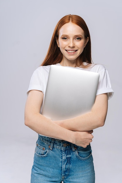 Charmante jonge vrouw student bedrijf laptopcomputer en kijken naar camera op geïsoleerde grijze achtergrond Mooie dame model met rood haar emotioneel gezichtsuitdrukkingen in studio kopie ruimte tonen