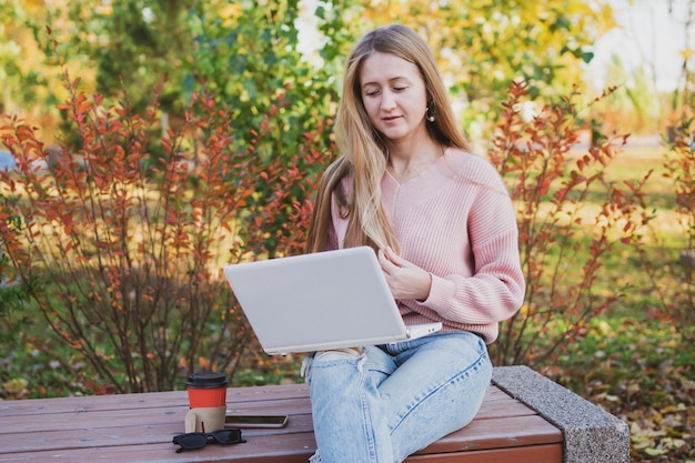 Charmante jonge vrouw werkt op laptop en drinkt koffie terwijl ze op de bank zit in een prachtige herfstpa