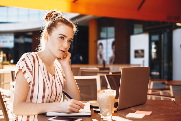 Charmante jonge zakenvrouw met rood haar en sproeten notities maken tijdens het drinken van koffie in een coffeeshop.