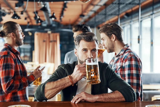 Charmante jongeman in vrijetijdskleding die bier drinkt terwijl hij tijd doorbrengt met vrienden in de kroeg