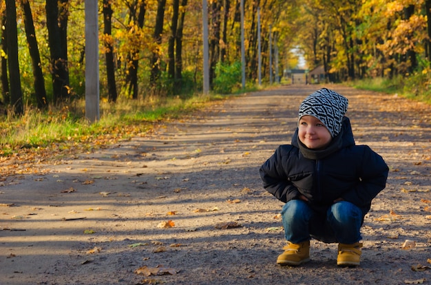 Charmante jongen van twee jaar in een blauwe jas en een bonte hoed ging in de herfst op het pad in het steegje van het park zitten