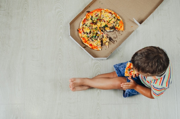 Charmante jongen zittend op de vloer pizza bovenaanzicht eten