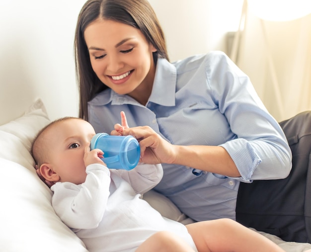 Charmante kleine baby drinkt terwijl hij thuis in de buurt van zijn zakelijke moeder op bed ligt