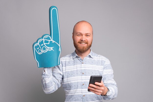 Charmante knappe kantoorman die wijst met een handschoen van schuim van een ventilator en een smartphone vasthoudt over een grijze achtergrond