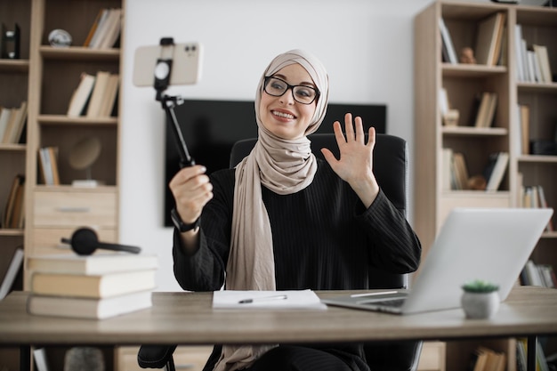 Charmante moslimvrouw in hijab praten en gebaren tijdens het opnemen van video op moderne telefoon op stick