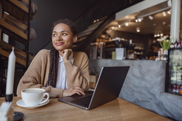 Charmante peinzende Afro-Amerikaanse vrouw in vrijetijdskleding genietend van werken op afstand op laptop en koffiepauze in gezellige cafetaria