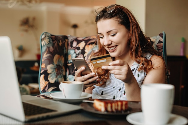 Charmante plus-size vrouwen met rood haar die een creditcard en een smartphone vasthouden die online transacties doet terwijl ze in een café zitten met een laptop.