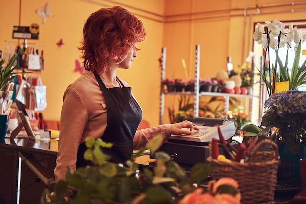 Charmante roodharige vrouwelijke bloemist die uniform draagt, werkt met een kassa in de bloemenwinkel.