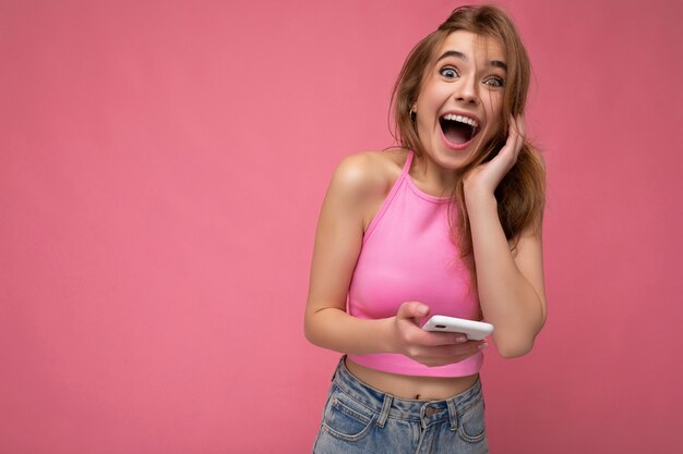 Charmante verbaasde positieve jonge blonde vrouw die roze top draagt ??die op roze achtergrond wordt geïsoleerd