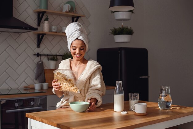 Charmante vrouw in een badjas en handdoek op haar hoofd bereidt een gezond ontbijt met vlokken en melk. Het meisje heeft 's ochtends een ontbijt in een stijlvolle, gezellige keuken.