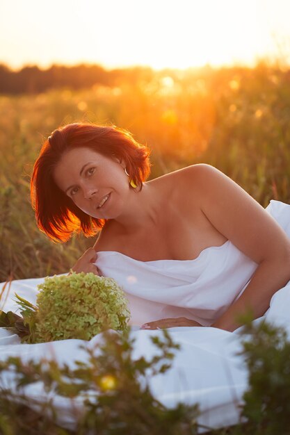 Foto charmante vrouw in wit bed in een veld bij zonsondergang in de zomer kijkend naar de camera