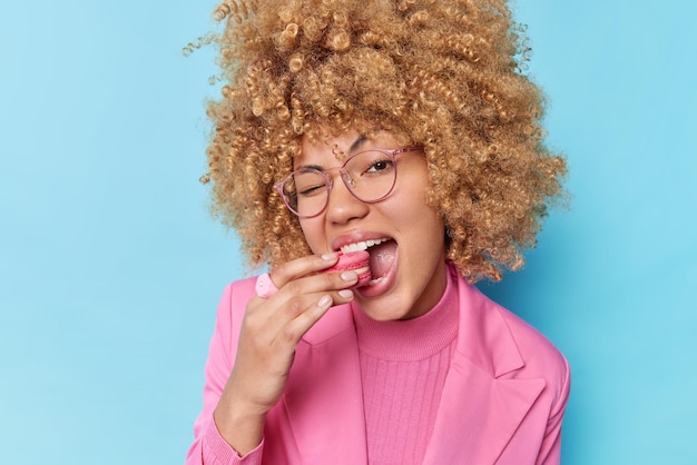 Charmante vrouw met krullend haar eet bitterkoekjes en knipoogt oog kijkt speels naar camera draagt transparante bril en pnk formele jas geïsoleerd op blauwe achtergrond Sweet tooth suikerachtig voedsel concept