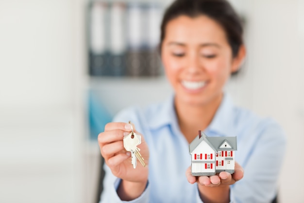 Charmante vrouw met sleutels en een miniatuur huis tijdens het kijken naar de camera