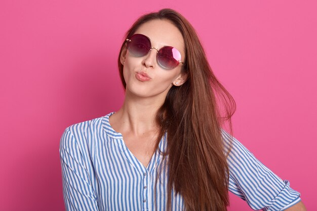 charmante vrouw met stijlvolle zonnebril en gestreept overhemd, houdt lippen afgerond, kus gebaar maken, model poseren geïsoleerd over roze muur