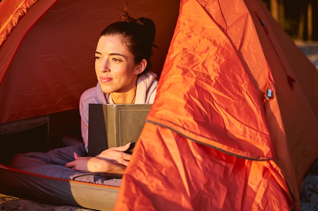 Charmante vrouwelijke reiziger met boek die wegkijkt en glimlacht terwijl hij op de buik ligt in een toeristische tent