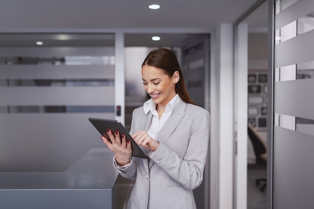 Charmante zakenvrouw in formele kleding en met een grote brede glimlach die tablet gebruikt terwijl hij in de hal op het werk staat.