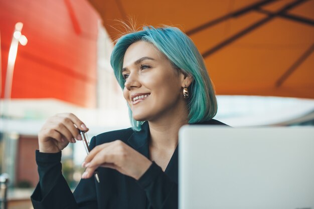 Charmante zakenvrouw met blauw haar, zittend in een restaurant en werken met een computer