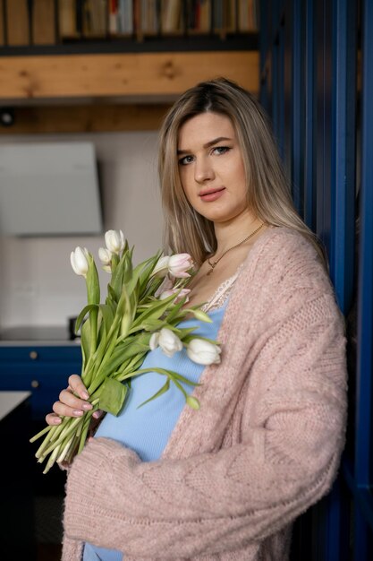 Charmante zwangere jonge vrouw in gebreid vest staat thuis met een boeket tulpen en zachtjes h