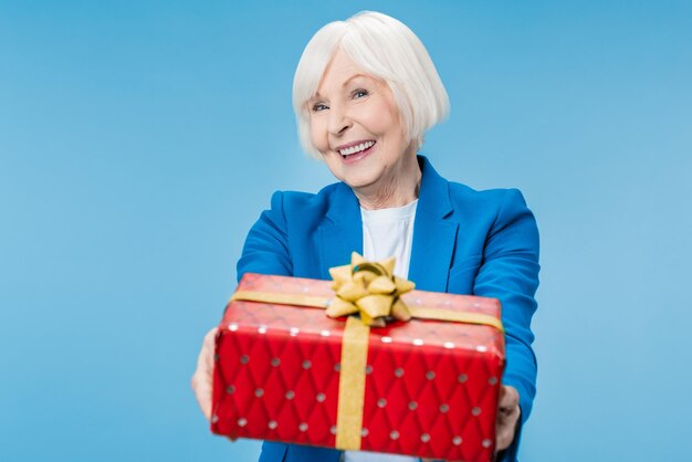 Charming aged woman giving gift box in red package with bow enjoying isolated on blue background