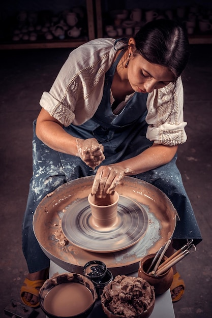 매력적인 소녀 조각가는 도공의 물레 위에서 점토로 작업하고 도구가 있는 테이블에서 작업합니다. 수공예품 생산.