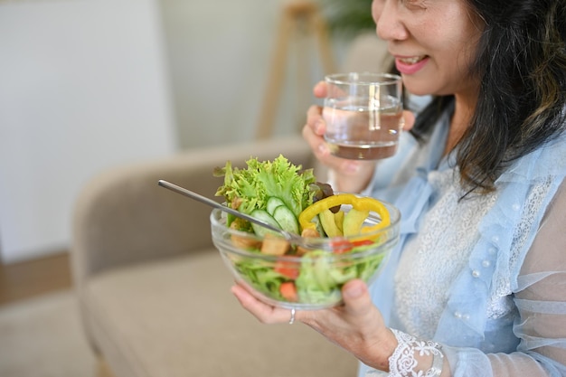 매력적인 아시아 60대 여성이 물을 홀짝이며 건강한 샐러드 그릇을 즐겨 먹습니다.