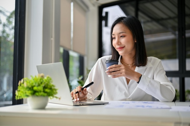 Charming Asian businesswoman sipping coffee while working on her financial project on laptop