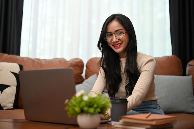 Charming Asian female in eyeglasses looking at the camera working from home using a laptop