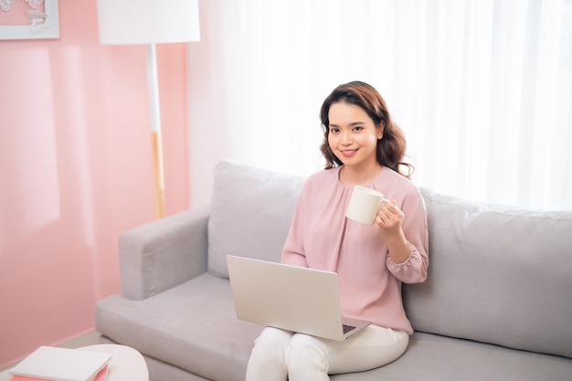 Charming Asian woman drinking coffee and using laptop when sitting on couch.