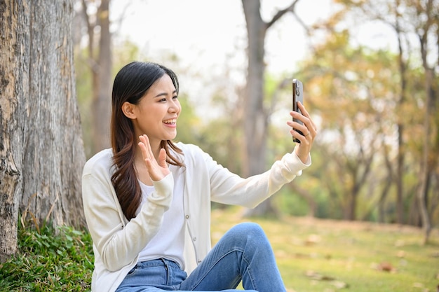 Charming Asian woman is talking on a video call with her friend while relaxing in the park