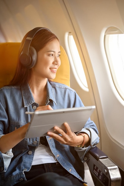 Charming asian woman passenger at window seat with headphones using portable tablet